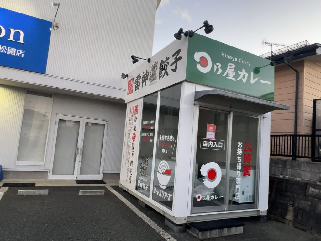 「工場直送 雷神餃子」「日乃屋カレー」「ヌードルツアーズ」の看板が掲出された無人販売店の外観